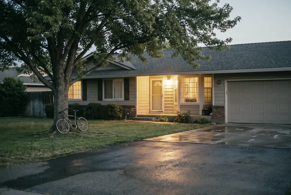 A single-storey Central Valley family home at dusk, its porch light and front windows glowing warm amber, a bicycle left on the lawn beneath a mature shade tree